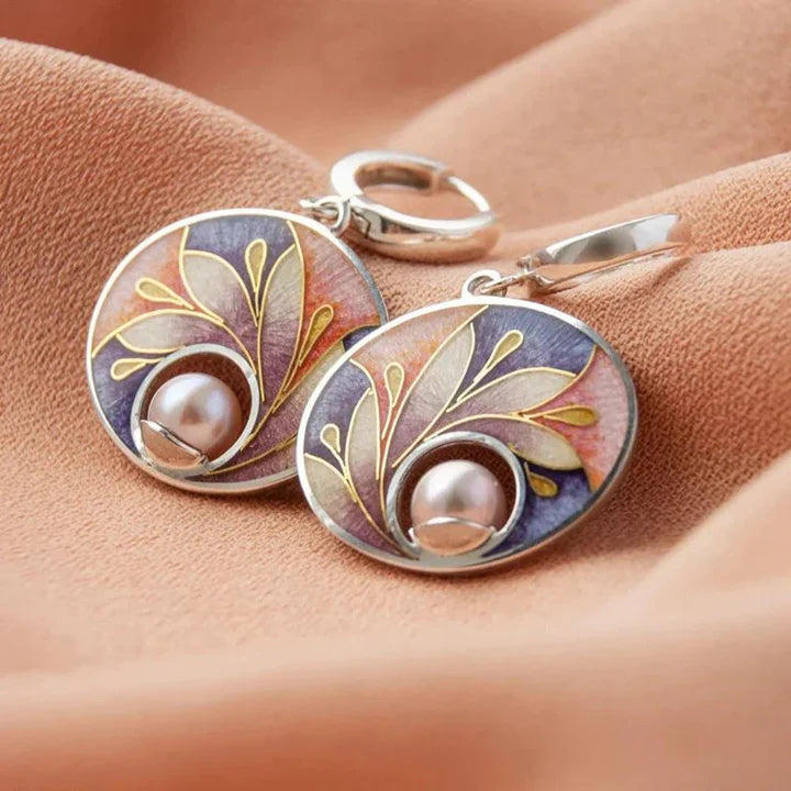These handcrafted Acrylic Earrings with Pearls feature round shapes, gold leaf patterns, purple and pink enamel accents, and a pearl at the base—all beautifully displayed on soft peach fabric. A perfect choice for those seeking high-quality jewelry.