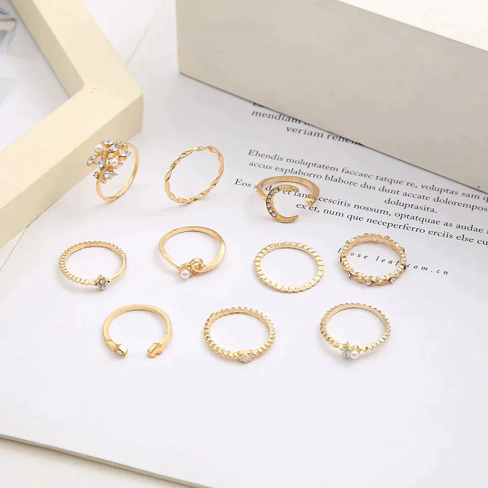 A 10 pack of Bohemian silver and gold plated rings with assorted designs, including twisted bands, pearls, and crystal accents, displayed on a white surface near an open book and a cream-colored frame.