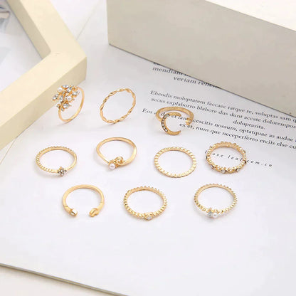 A 10 pack of Bohemian silver and gold plated rings with assorted designs, including twisted bands, pearls, and crystal accents, displayed on a white surface near an open book and a cream-colored frame.