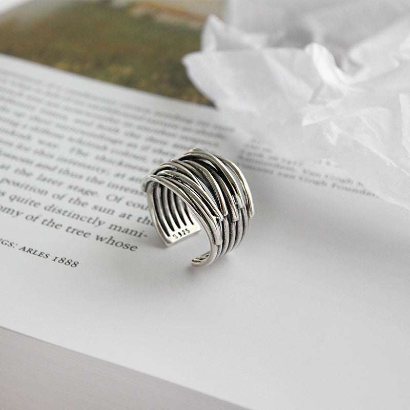 The 925 Sterling Silver Adjustable Layered Ring, featuring overlapping bands for a textured look, is displayed on an open book with crumpled white tissue paper in the background.