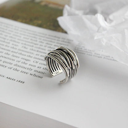 The 925 Sterling Silver Adjustable Layered Ring, featuring overlapping bands for a textured look, is displayed on an open book with crumpled white tissue paper in the background.
