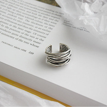 The 925 Sterling Silver Adjustable Layered Ring from our handcrafted jewelry collection rests on an open book with white pages, beside a crumpled piece of tissue paper. Some text on the book page is partially visible.