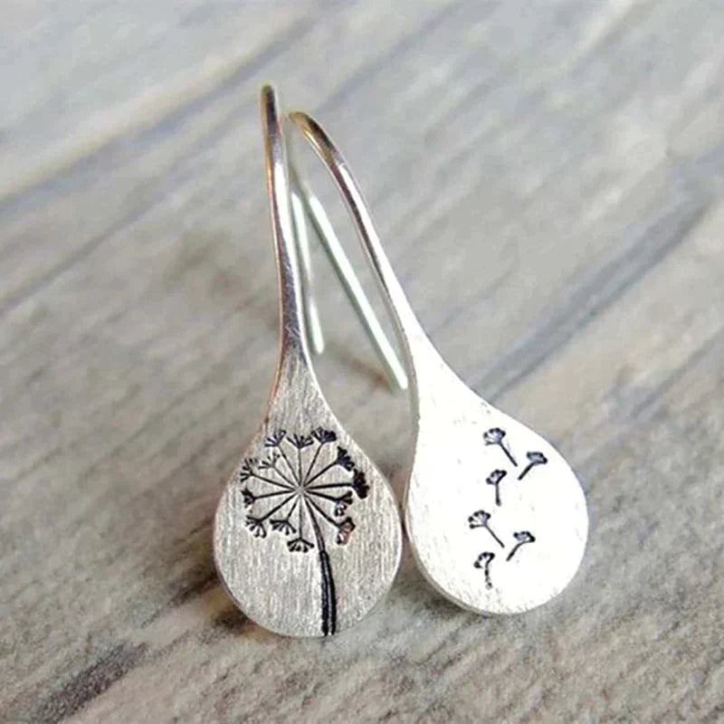 The 925 Sterling Silver Dandelion Leaf Earrings are handcrafted and engraved with dandelion designs—one earring shows a full dandelion, the other has seeds blowing away. This elegant pair is displayed on a light wooden surface.