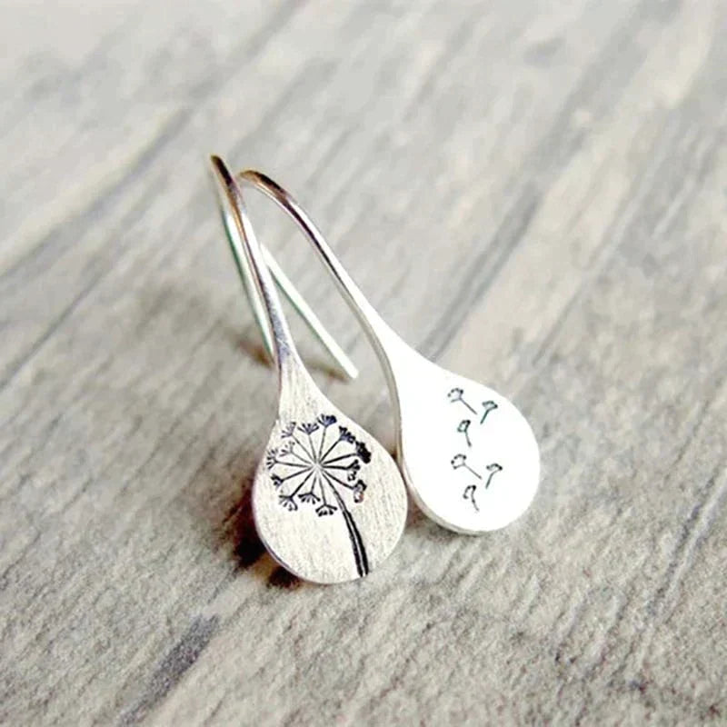 The 925 Sterling Silver Dandelion Leaf Earrings—a mismatched pair featuring a dandelion flower and blowing seeds—capture the beauty of handcrafted jewelry, elegantly presented on a wooden surface.