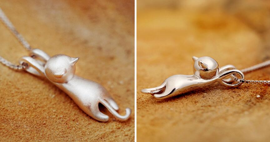 The 925 Sterling Silver Playful Kitty Necklace features a smooth, minimalist pendant shaped like a stretching cat with outstretched front legs, shown from two angles against a warm brown background—a beautiful handcrafted jewelry piece.